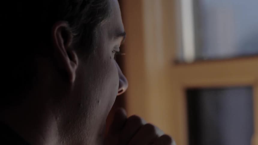 A young man nervously moves his hands about his face as he sits in front of a window