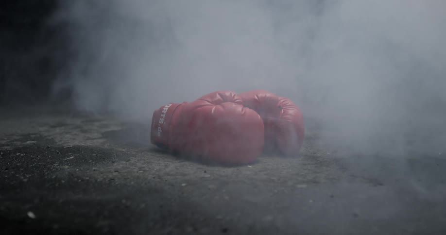 Smoke blows over and around a pair of red boxing gloves on the ground