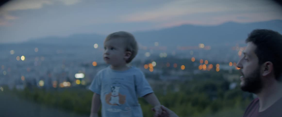 A male toddler stands outside with his father on a hilltop above a city below
