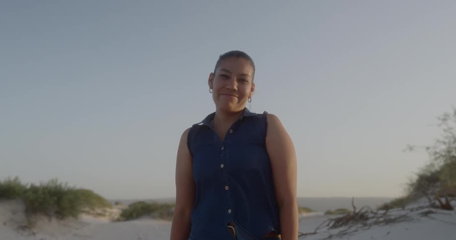 Latino woman looking at camera standing on beach next to the ocean