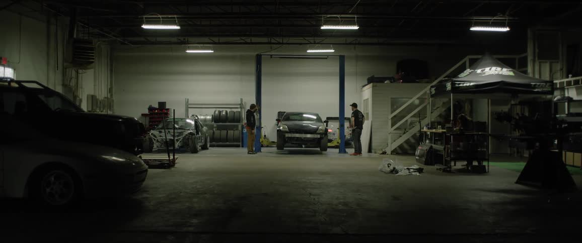 Several men stand in a garage and watch a car being jacked up