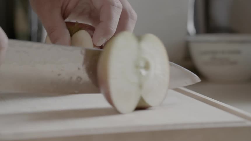 Male slices an apple that is sitting on a cutting board down the middle with a large sharp knife