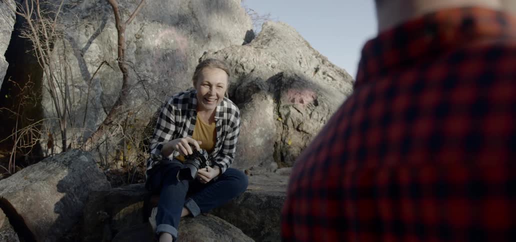 A woman sits on a rock across from a man and takes a photo of him