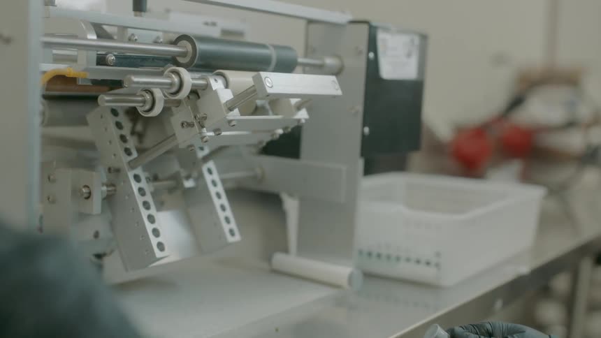 A worker wearing glove places tube after tube into a machine that applies a label to each one