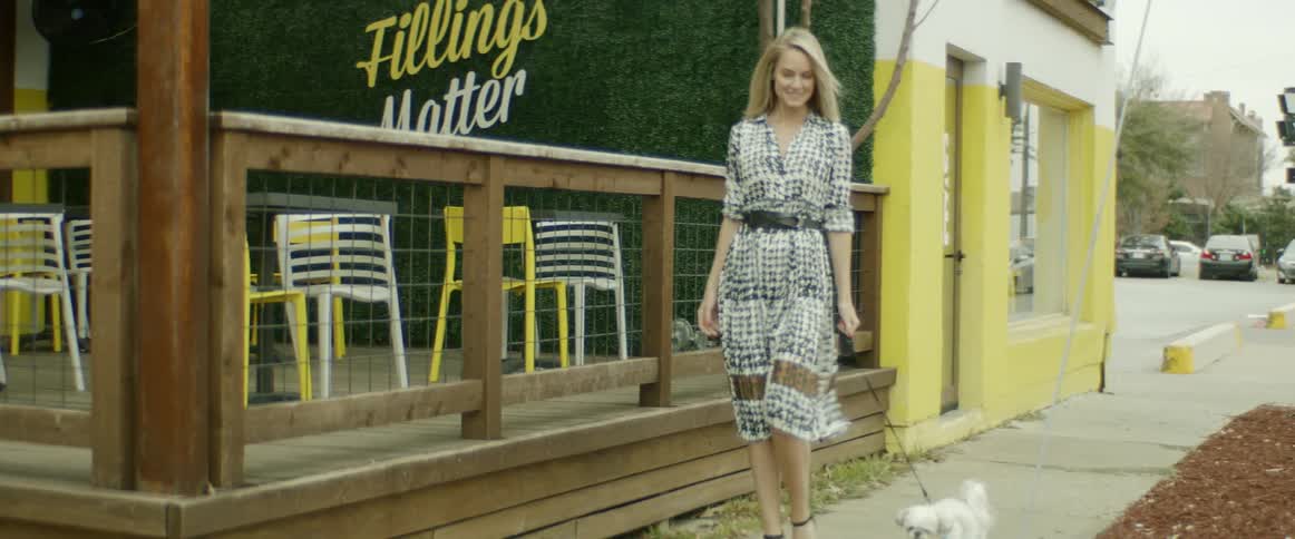A white female in a dress and high heels is walking a small white dog on the sidewalk next to a business with outdoor seating