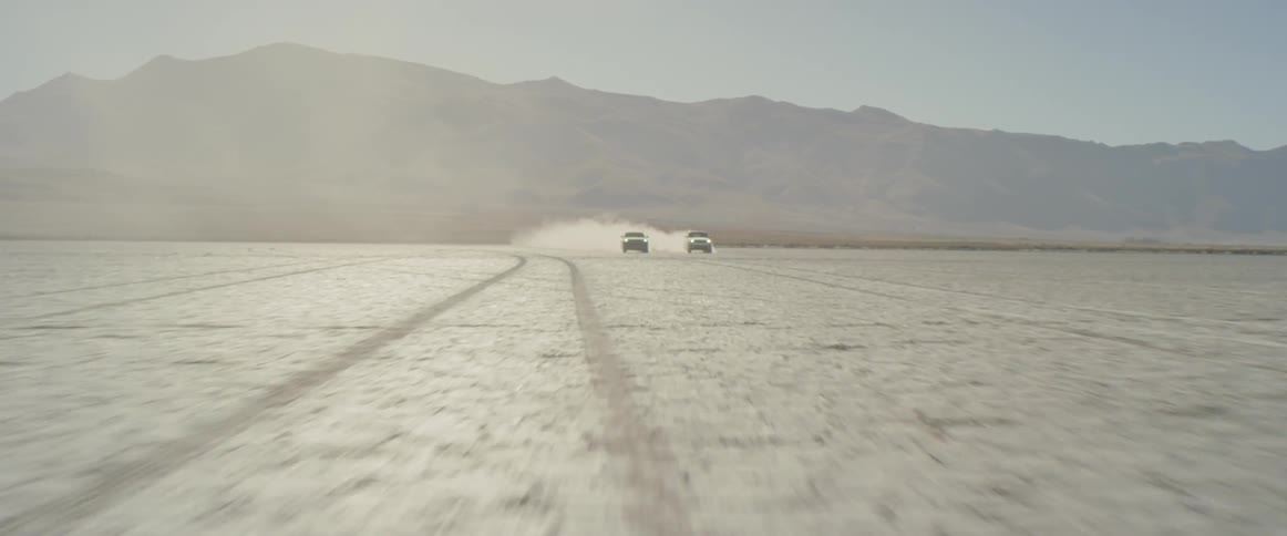 Two vehicles race across a desert away from the mountains