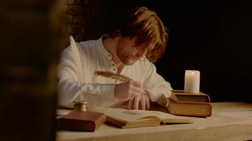 Man sitting at a table writing with a feather tip ink pen by candle light
