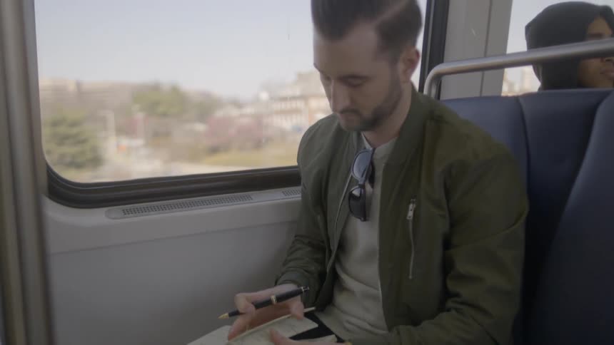 A young man sits on a bus thumbing through his notebook