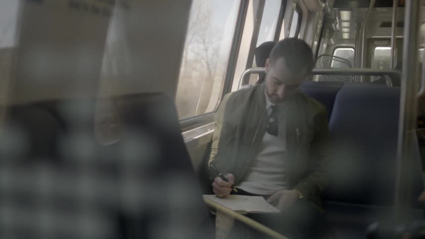 A man sitting on a train while writing in a notebook