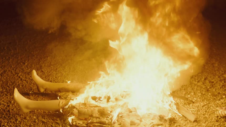 The legs of a mannequin stick out from a smoky flaming fire