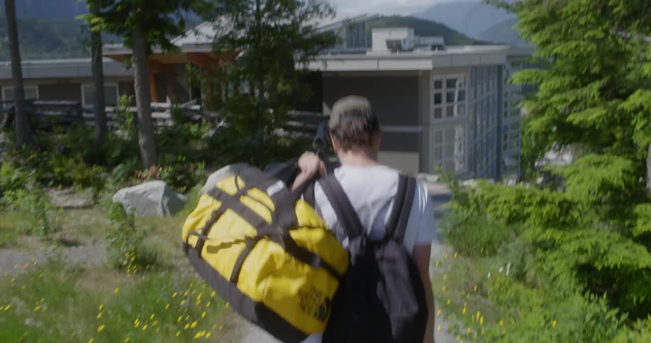 A white man carrying heavy bags and wearing a backpack walks on a path to a house