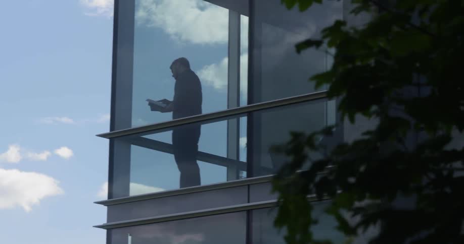 A man stands in a corner of a building with floor to ceiling windows and talks on the phone