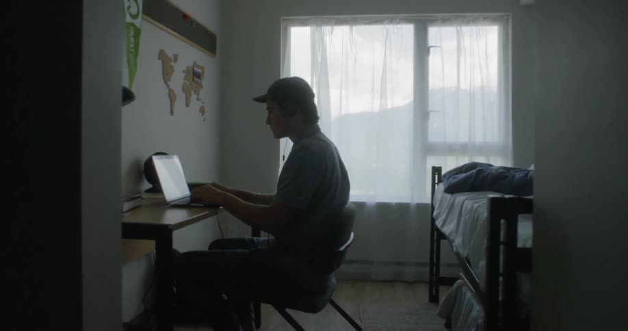 Teenager sitting at desk playing music on his laptop in his room