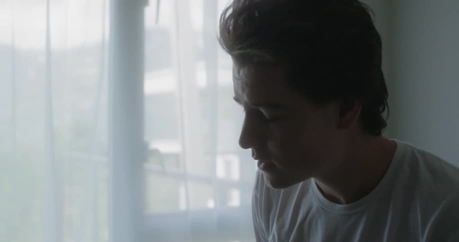 A young man takes off his hat looks out the window and rubs his hands over his face and through his hair