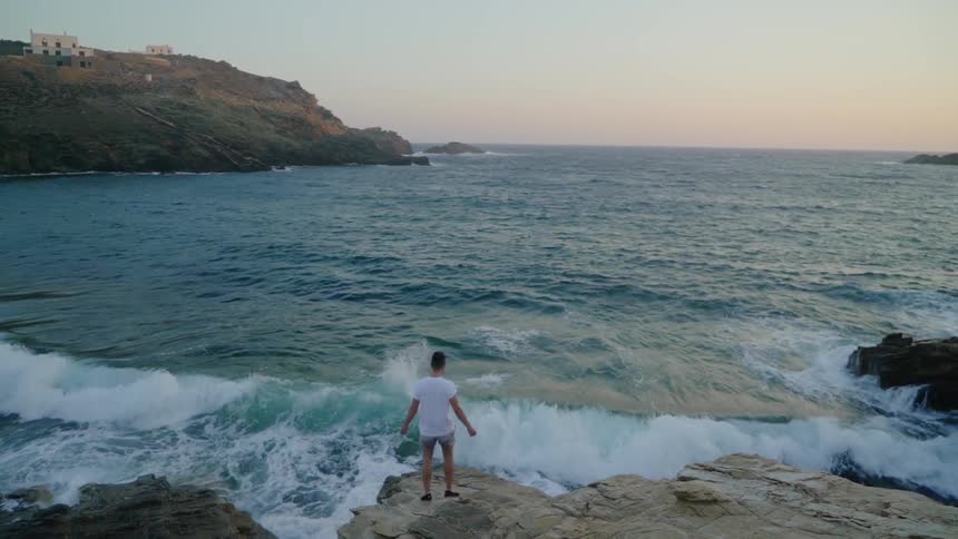 Man stands on a rock and raises his arms as he looks out at the ocean