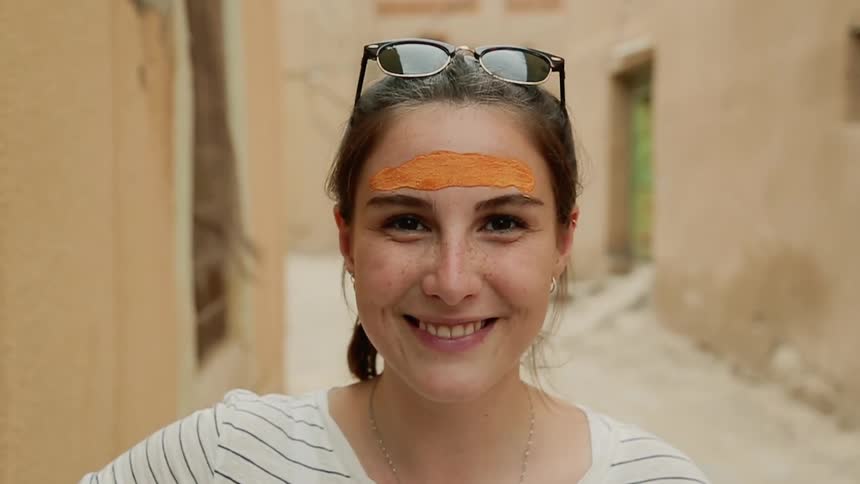 A young woman with paint on her forehead smiles and poses for a camera