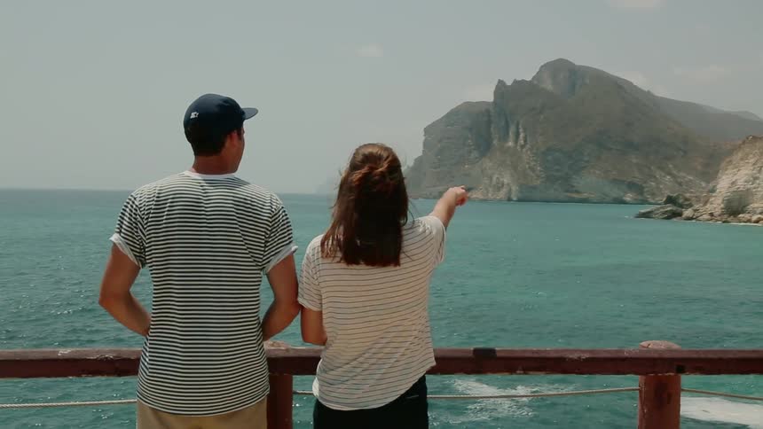 Man and woman stand near a railing as they look out at the ocean and mountains