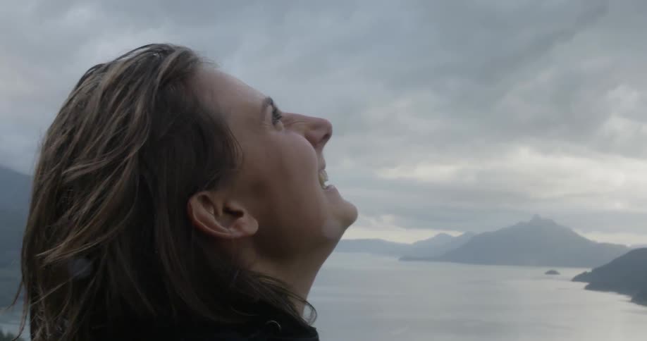 Woman laughs as she stands near a lake and looks up at the cloudy sky