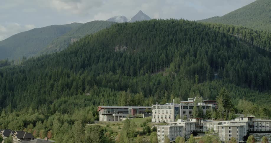 A number of modern buildings stand out amidst a densely forest area at the base of a range of hills and mountains