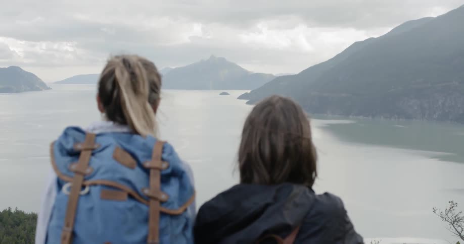 Two females wearing backpacks are lookingout over a lake thats surrounded by mountains on a cloudy day