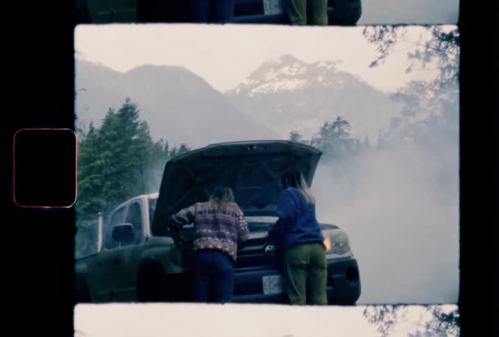 Two females are looking under the hood of a smoking truck with mountains in the background