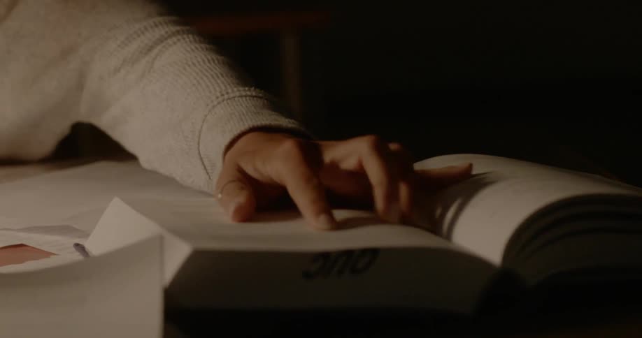 A young woman hurriedly reviews her books and notes as she sits at a desk in a dimly lit room