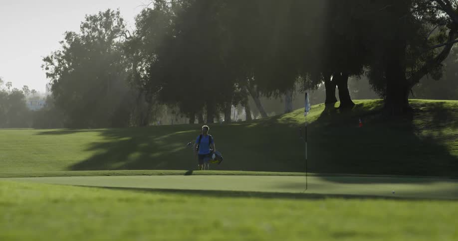 A white male golfer is carrying his golf bag and clubs to the putting green on a golf course