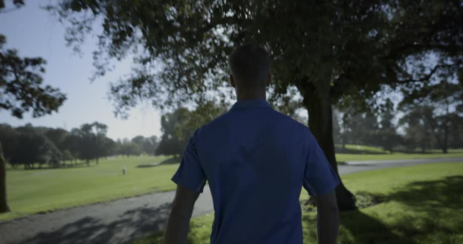 Golfer walks through golf course through trees to paved road