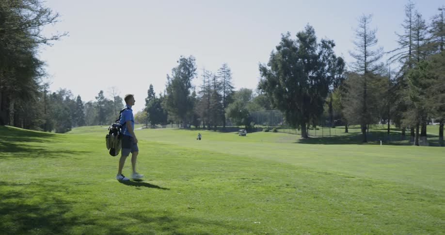 A man with a golf bag strapped over his shoulder walks swiftly across the green grass of a golf course