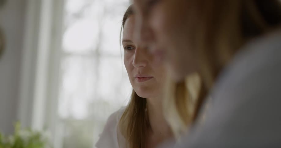 Two women sitting at the table look at the laptop and talk to each other in the room