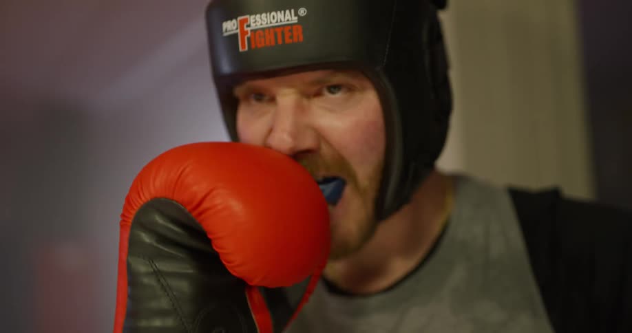 A man wearing boxing gloves and headgear place a mouth guard in his mouth in preparation for sparring