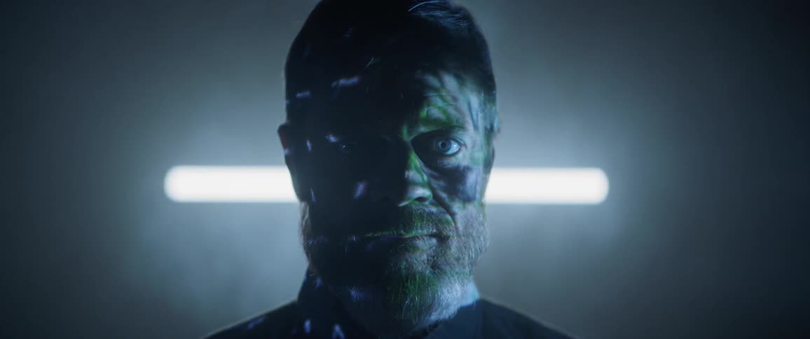 A man stares into the camera while lights and shapes are projected onto his body and face