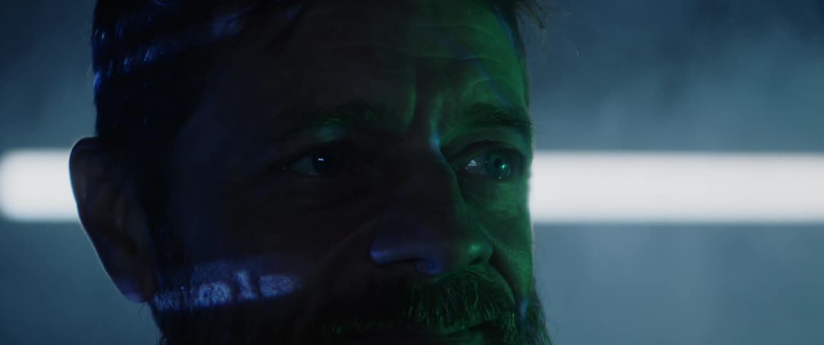A bearded man stares off in the distance as computerized text is projected onto his face in a darkened and foggy room