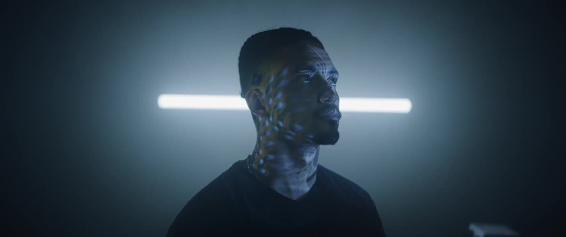 A black man sits as lights from a screen reflect on him