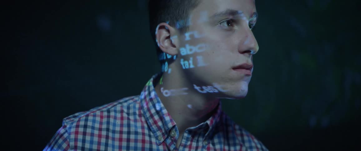 A teenager sits in front of a projector screen and has film and text flashed on his face