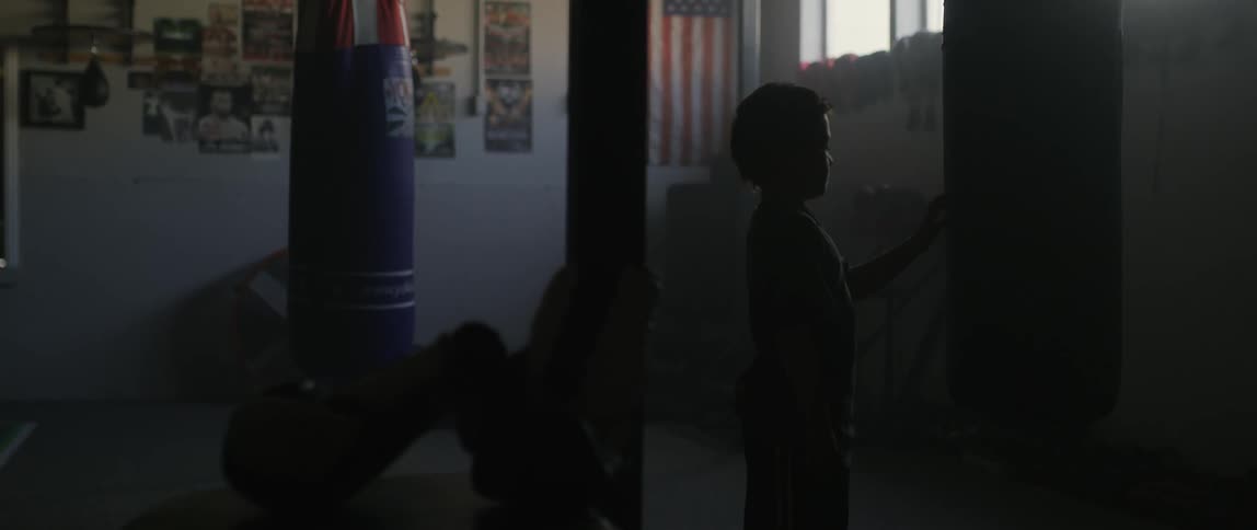 A child walks in the space between a set of boxing gloves and a punching bag in a dark gym