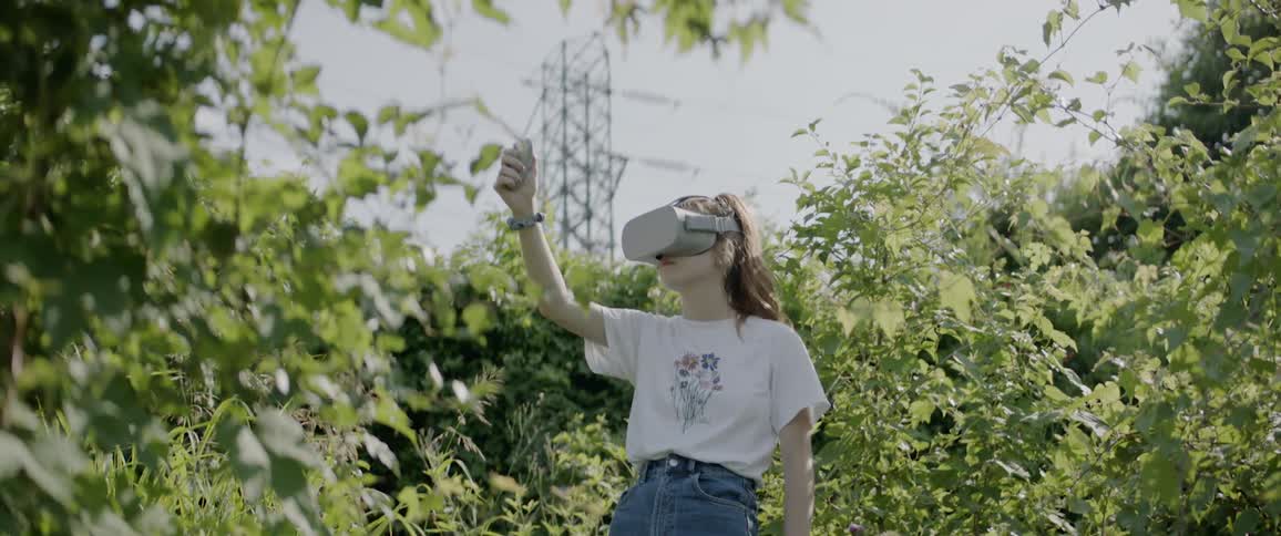 A young female stands outdoors amidst thick vegetation while wearing virtual reality goggle and waving a controller in her hand