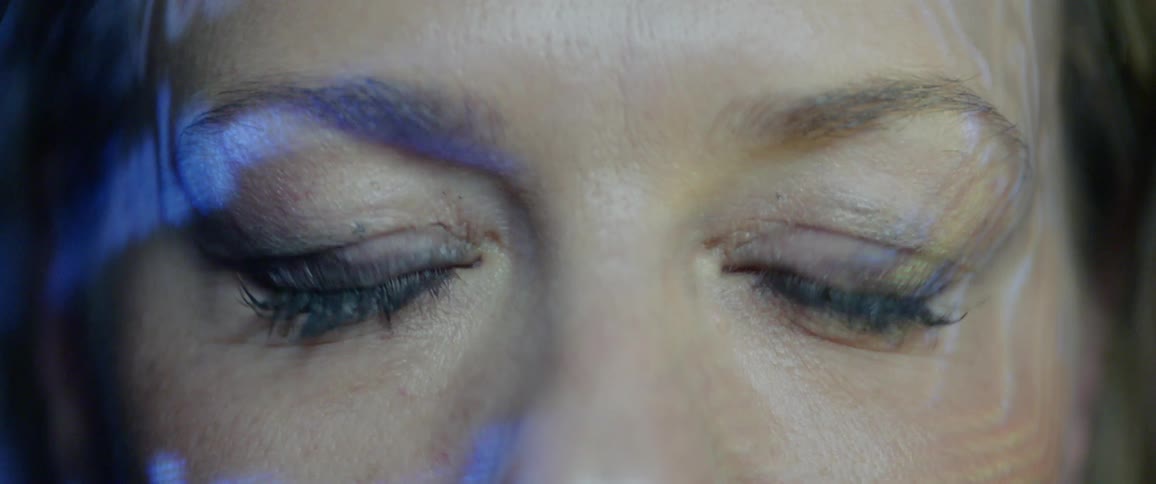 A woman blinks her blue eyes while lights and text flash across her face