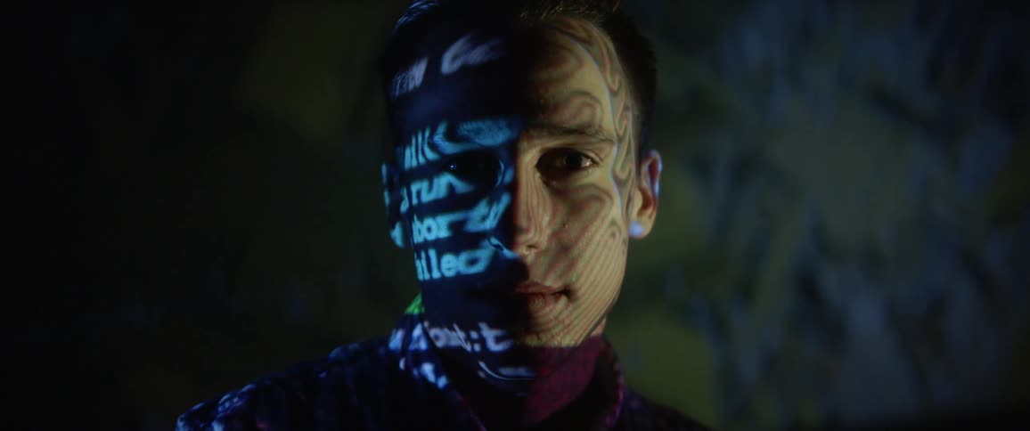 A man sits in a darkened room as blue and green computerized text is projected onto his face