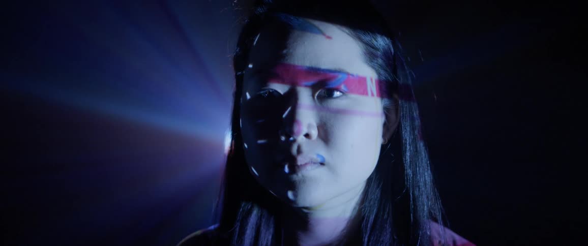 An asian woman standing in a dark roll as images are projected on her face and multicolored light shines behind her