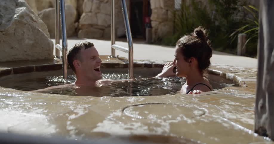 A white couple is talking and smiling while they soak in an outdoor hot tub