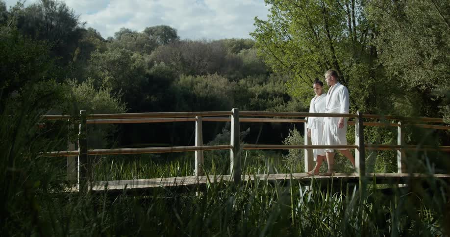A man and woman each wear white bathrobes as they walk outdoors over a small bridge