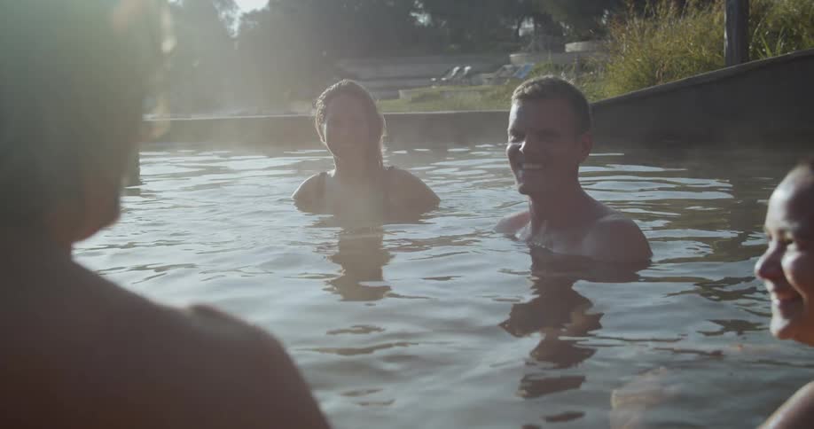 A group of people are all sitting in a large outdoor spa while smiling and conversing
