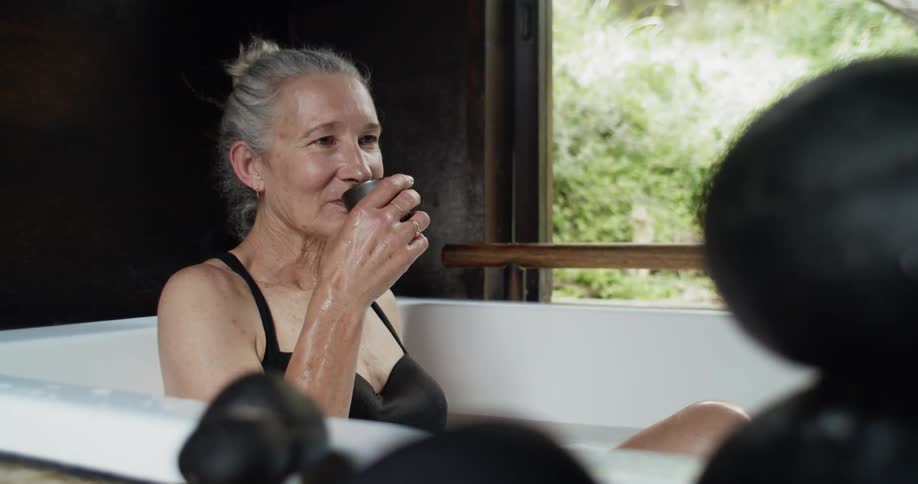 Older woman drinks from cup while sitting in bathtub next to window