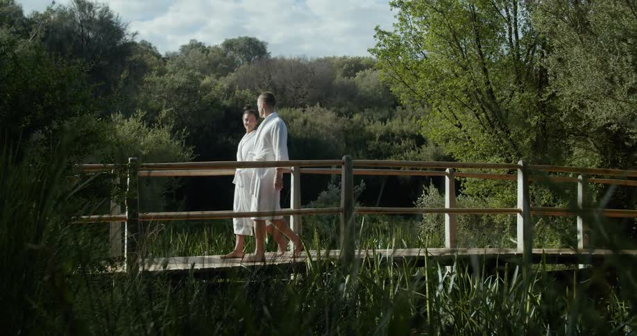 A couple in bathing robes walking along a wooden bridge surrounded by marshland and trees