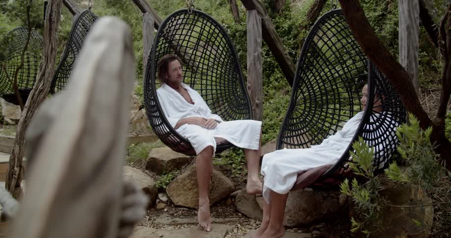 A couple wearing white bathrobes lounge in hanging basket chairs outdoors while chatting