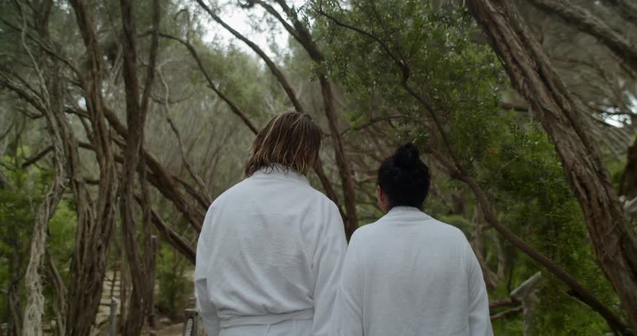 A couple wearing white bathrobes holds hands as they walk on a wooded trail