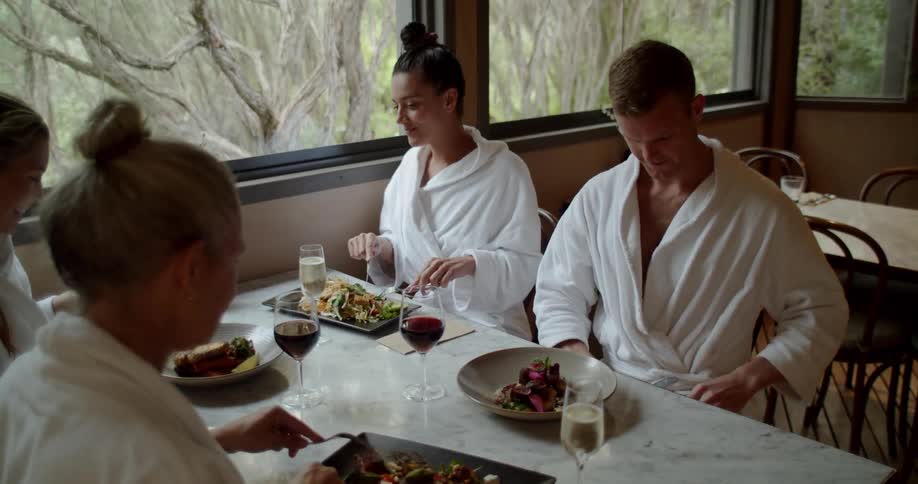 A group of friends dressed in terry cloth robes sit at a dining table talking and sharing a meal