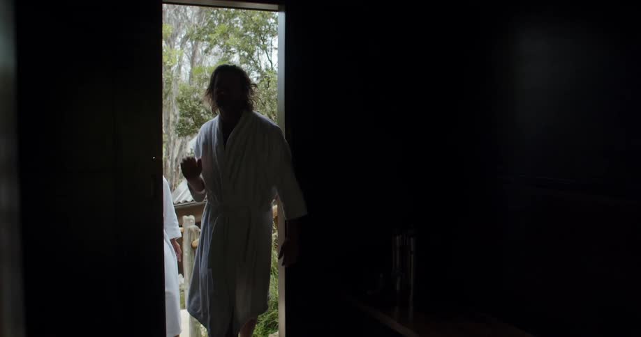 A man in a white bathrobe opens the door to a darkened room and then he and his female companion step inside