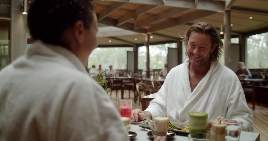 A man smiles and laughs with his female companion as the two sit at dining table in a dining area while the two wear white bath robes